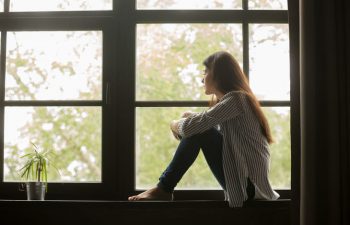Depressed woman sitting on the windowsill inside the house and looking through the window