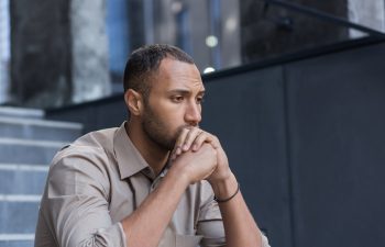 A man sits outdoors on stairs with his hands clasped in front of his mouth, looking thoughtful and serious.